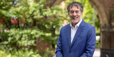 Alastair Da Costa, Chair of the University of Leeds Council, smiling outdoors in a blue suit, with greenery and historic university architecture in the background
